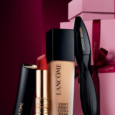An elegant still life of Lancome's L'Absolu Rouge matte lipstick, Teint Idole Ultra Wear foundation, and Hypnose Drama mascara set against a luxurious dark red holiday background.