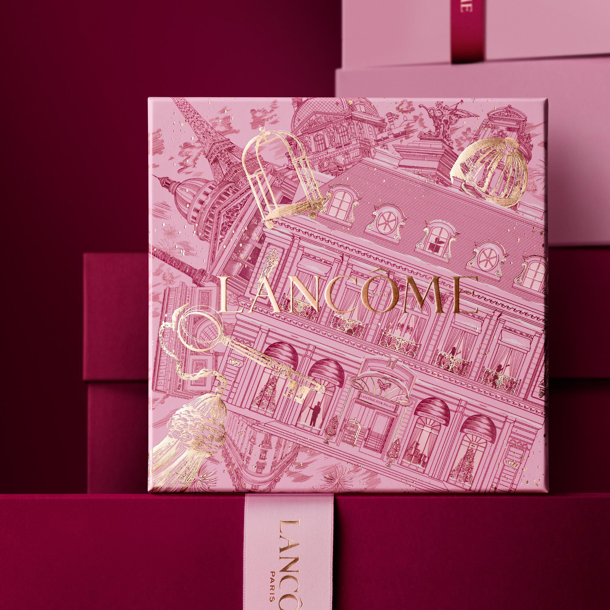 Image of pink Lancome holiday gift box standing in front of a pile of pink and dark red gift boxes, set against a dark red background.