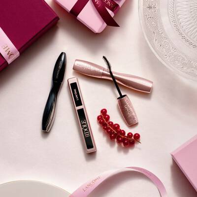 A flat-lay arrangement of Lancome makeup products, featuring mascara and lip gloss, alongside pink gift boxes, satin ribbons, and a sprig of red currants.