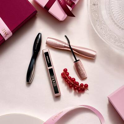 A flat-lay arrangement of Lancome makeup products, featuring mascara and lip gloss, alongside pink gift boxes, satin ribbons, and a sprig of red currants.