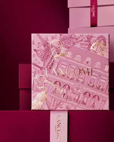 Image of pink Lancome holiday gift box standing in front of a pile of pink and dark red gift boxes, set against a dark red background.