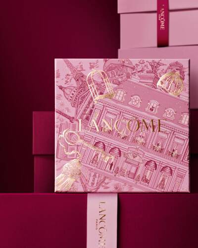 Image of pink Lancome holiday gift box standing in front of a pile of pink and dark red gift boxes, set against a dark red background.