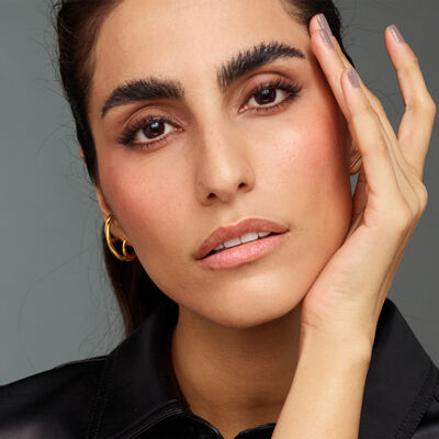 A close-up portrait of a woman gently touching her face, with expressive eyebrows and a natural makeup look, set against a neutral background.