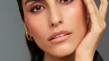 A close-up portrait of a woman gently touching her face, with expressive eyebrows and a natural makeup look, set against a neutral background.