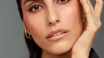 A close-up portrait of a woman gently touching her face, with expressive eyebrows and a natural makeup look, set against a neutral background.