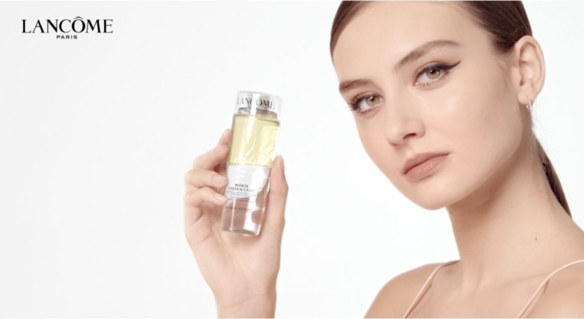 A model holds a bottle of Lancome skincare product, showcasing it with a soft smile against a light background. The Lancome logo is displayed in the top left corner.