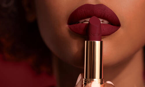 Close-up of a person holding a tube of deep red lipstick near their lips, which are painted in the same color, showcasing a matte finish.