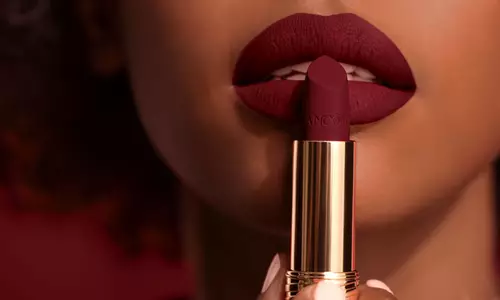 Close-up of a person holding a tube of deep red lipstick near their lips, which are painted in the same color, showcasing a matte finish.