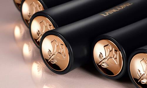 A close-up image of several black cosmetic tubes with gold caps, featuring an embossed floral design and the word "Lancome" in gold lettering.