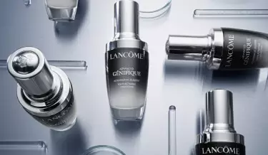 An arrangement of Lancome skincare products, featuring sleek, silver and black bottles of Advanced Genifique serum, set against a reflective metallic background.