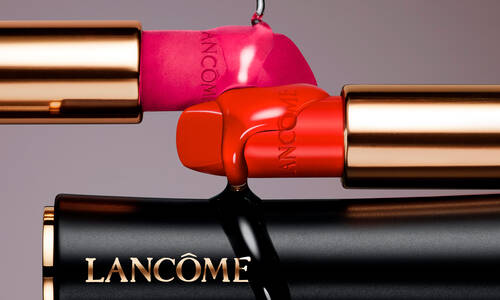 Three lipstick tubes in shades of pink and red are artistically arranged. The tubes have gold accents and the Lancome logo prominently displayed. The background is a soft gradient.