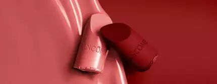 A close-up image featuring two lipsticks, one light pink and one deep red, against a smooth, red background. The lipsticks are partially unwrapped, displaying their sleek designs with the brand name "Lancome" visible on the casing.