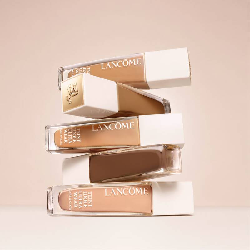 A stack of four bottles of Lancome foundation, each with a simple white cap and labeled "Teint Idole Ultra Wear." The bottles feature varying shades of foundation against a soft beige background.