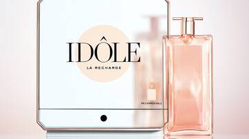 A sleek, modern perfume bottle with a soft pink liquid is displayed next to a reflective square container labeled "IDOLE LA RECHARGE." The design features a minimalist aesthetic with elegant lines.