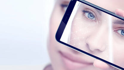 A close-up of a person holding a smartphone to their face, displaying a digital skin analysis overlay on the screen. The individual has striking blue eyes and a neutral expression.