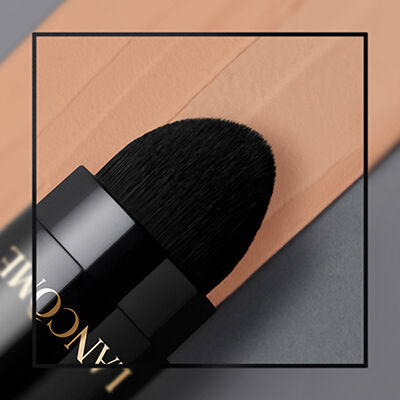 Close-up of a makeup brush with a soft, black bristle tip resting next to a swatch of peach-toned foundation on a gray background.