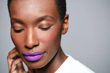 A close-up portrait of a woman with short hair, wearing vibrant purple lipstick and a soft expression, against a neutral gray background.