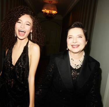 Two women, one with curly hair laughing and another smiling, both dressed in formal dark attire, are pictured in a hallway.