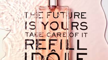 A transparent bottle of perfume sits on a smooth surface, surrounded by a splash of water. The words "THE FUTURE IS YOURS TAKE CARE OF IT REFILL" are printed beneath the bottle in bold, contrasting letters.