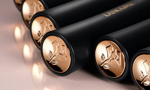 Close-up of black cylindrical tubes with gold caps, featuring an embossed floral design, arranged in a row. The branding "Lancome" is visible on the tubes. The surface reflects light, adding a glossy effect.