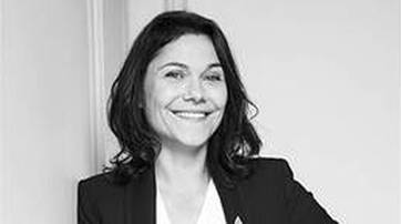 A smiling woman with dark, wavy hair, wearing a black blazer over a white top, stands against a light-colored wall.