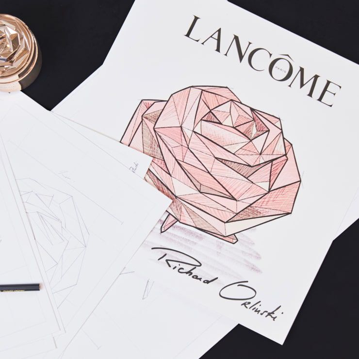 An artistic sketch of a geometric rose, labeled "Lancome" at the top. The sketch features shading in pink tones and includes the signature "Richard Orlinski" at the bottom. Surrounding the main image are several blank sheets of paper and a pen.