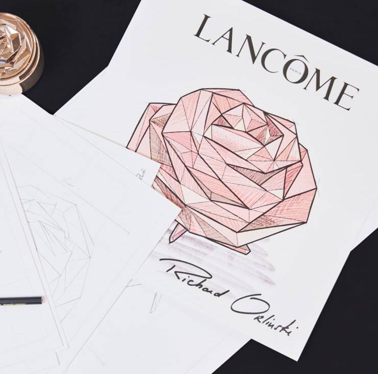 An artistic sketch of a geometric rose, labeled "Lancome" at the top. The sketch features shading in pink tones and includes the signature "Richard Orlinski" at the bottom. Surrounding the main image are several blank sheets of paper and a pen.