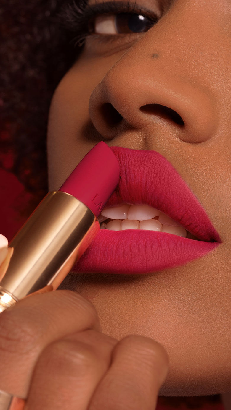 Close-up of a person applying dark pink lipstick to their lips, showcasing a smooth, matte finish. The background features a subtle red hue.