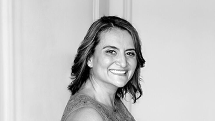 A woman with shoulder-length hair smiles at the camera, wearing a stylish dress. The image is presented in black and white.