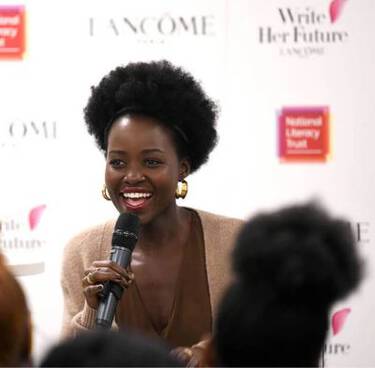 Lupita Nyong'o smiles while speaking into a microphone, with "LANCOME" and "Write Her Future" logos in the background.