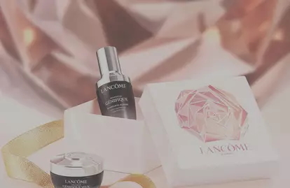 A stylish gift box featuring two Lancome skincare products: a serum and a cream, set against a sparkling, abstract background. There is a gold ribbon adorning the scene.