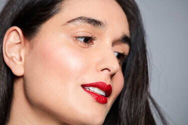 A close-up portrait of a woman with long, dark hair, smiling softly. She has a light makeup look with shimmery skin and bold red lipstick, set against a neutral gray background.