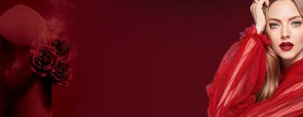 A glamorous woman in a flowing red outfit poses next to a dark red cylindrical container adorned with roses, set against a deep red background.