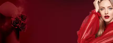 A glamorous woman in a flowing red outfit poses next to a dark red cylindrical container adorned with roses, set against a deep red background.