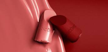 Close-up of two lipstick tubes in shades of pink and red, with a smooth, glossy red background and the Lancome logo visible on the tubes.