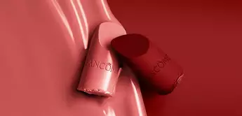 Close-up of two lipstick tubes in shades of pink and red, with a smooth, glossy red background and the Lancome logo visible on the tubes.