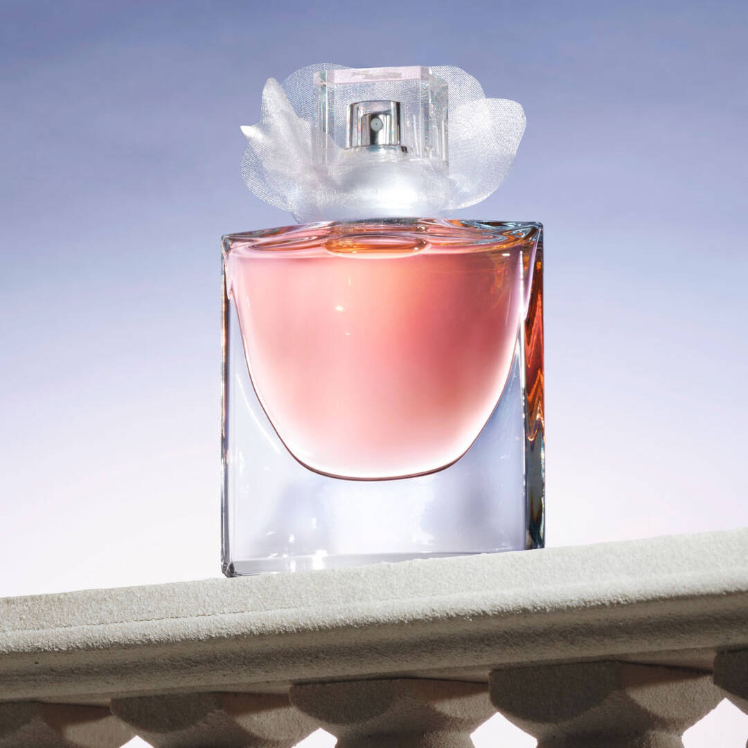 A clear glass perfume bottle with a soft pink liquid inside, topped with a delicate white flower-shaped cap, is displayed against a gradient background.