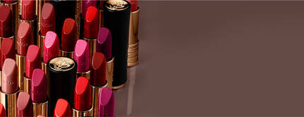 A variety of lipsticks in different shades of red, pink, and coral, neatly arranged with some of the caps visible on a brown background.