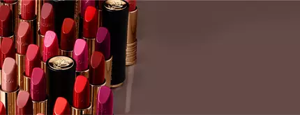 A variety of lipsticks in different shades of red, pink, and coral, neatly arranged with some of the caps visible on a brown background.