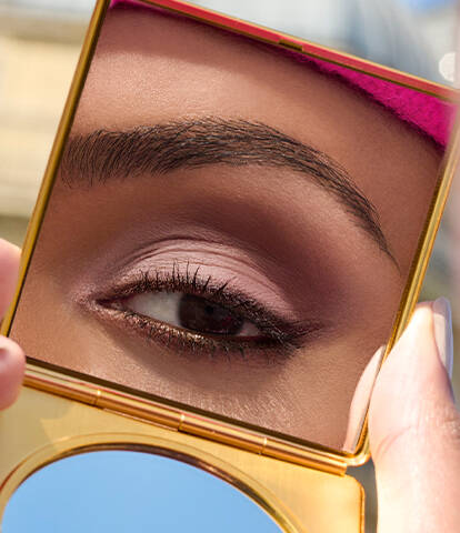 A close-up of an eye reflected in a compact mirror, featuring soft pink eyeshadow and perfectly groomed eyebrows. The background appears blurred, emphasizing the eye's details.