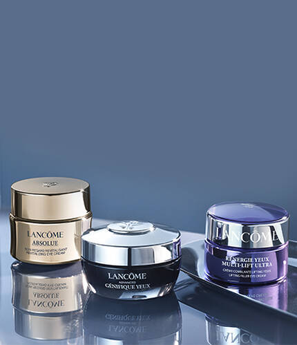 A close-up of three jars of Lancome skincare products displayed elegantly on a reflective surface, featuring luxurious gold, silver, and purple packaging.