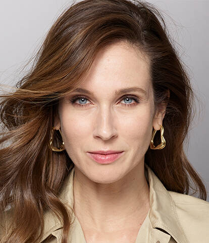 A woman with long, flowing hair and a neutral expression poses against a gradient background, wearing a light-colored jacket and statement earrings.