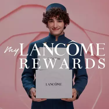 A smiling man in a blue bellhop uniform holds two white Lancome rewards boxes, with the text "my LANCOME REWARDS" overlaid on the image.