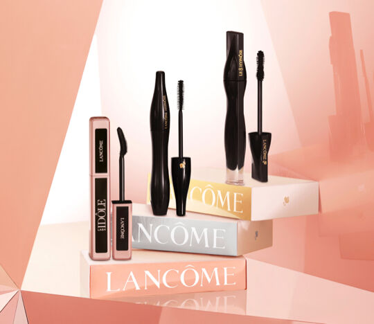 A display of various Lancome mascara products arranged on colorful boxes, highlighting their sleek black packaging against a soft, geometric background.
