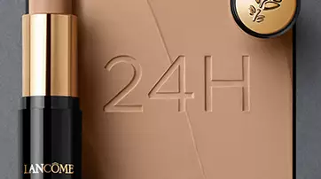 A Lancome foundation stick with a soft applicator next to a small compact, displayed on a background with swirls of foundation. The number "24H" is embossed in the surface.