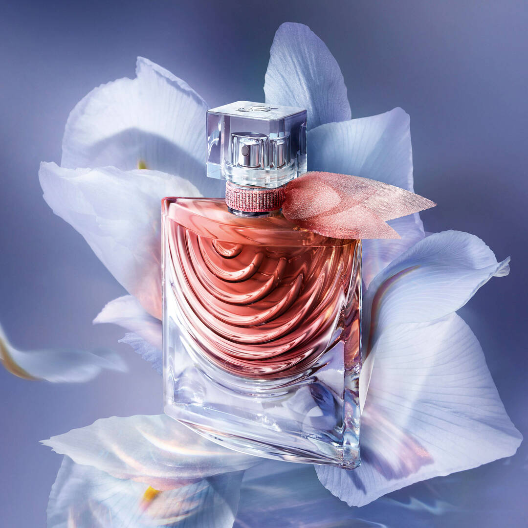A bottle of perfume with a curved design and a pink hue, surrounded by delicate flower petals against a soft, blurred background.