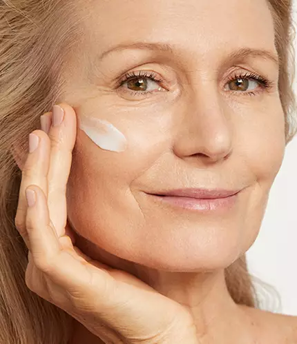 A woman with light hair gently applying cream to her cheek, smiling softly at the camera.