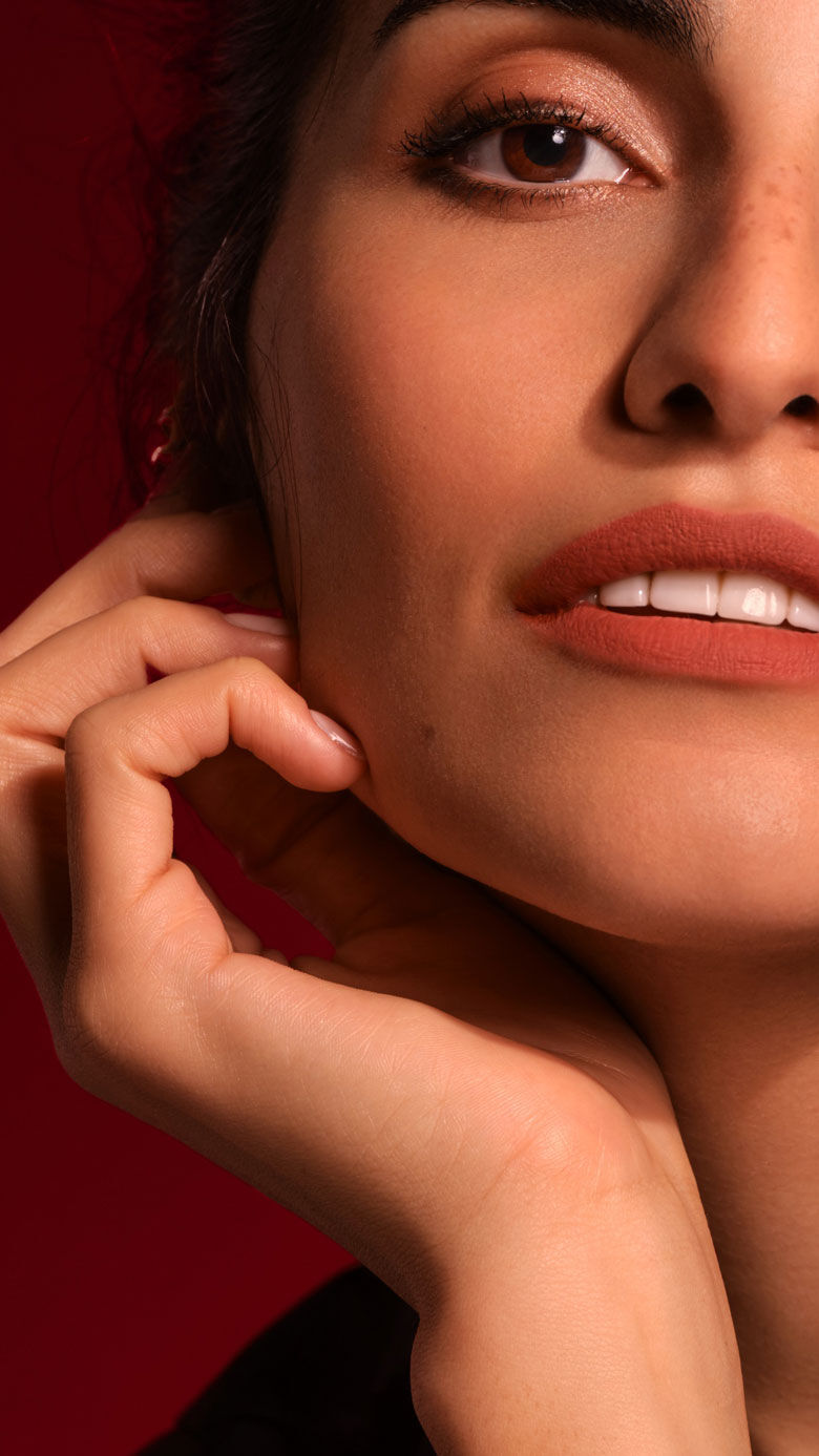 A close-up portrait of a woman with soft makeup, highlighting her features, and her hand gently touching her face against a dark red background.