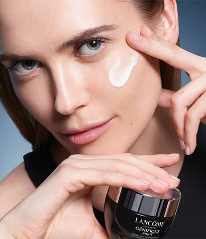 A woman applies cream to her face while holding a jar of skincare product, posing confidently against a gradient blue background.