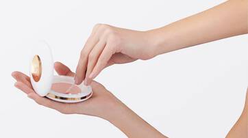 A hand holding a compact makeup case while another hand reaches inside to take some product. The compact has a sleek white design with a gold accent.